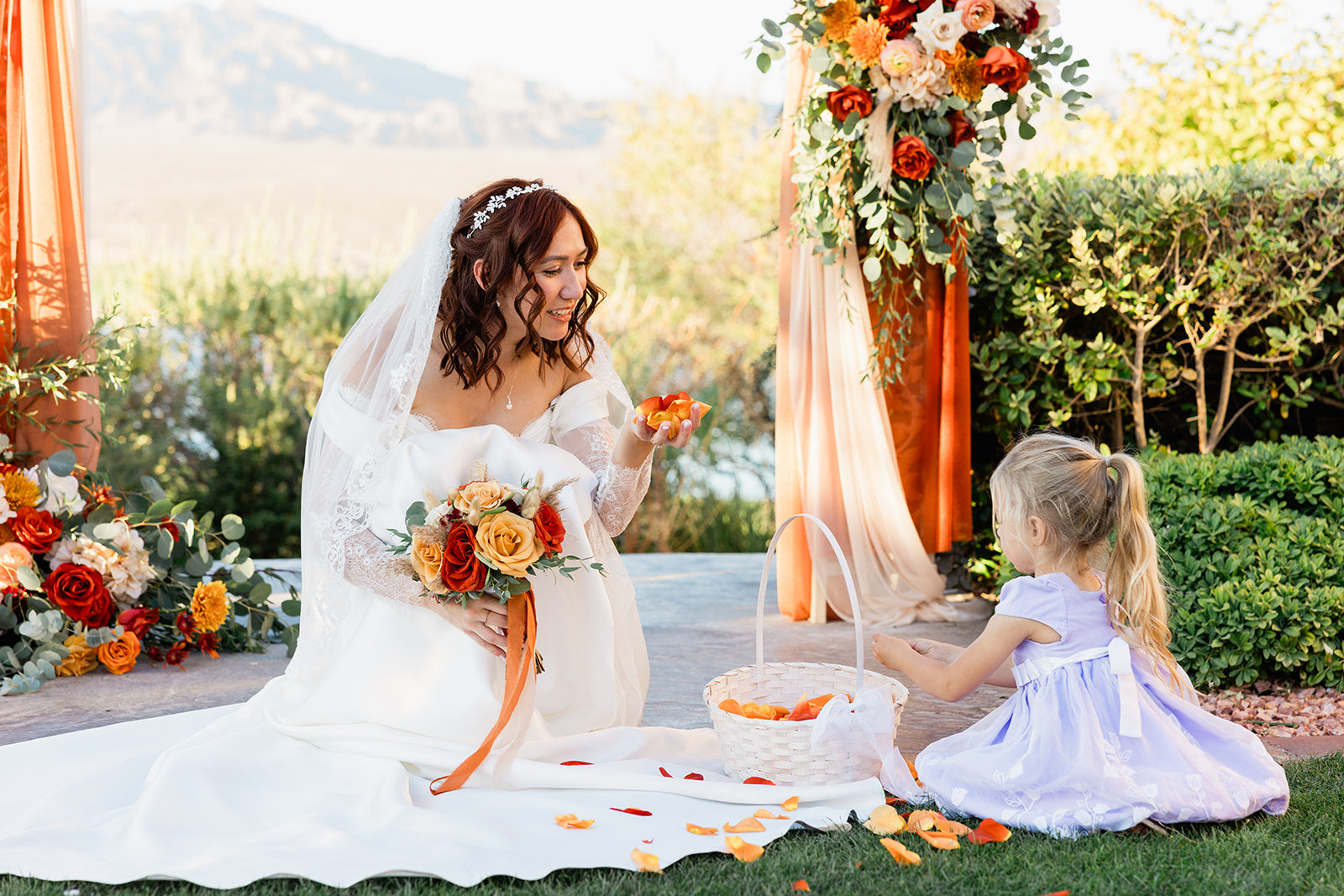 Bride in a white wedding dress with a veil, holding a bouquet, interacting with a young girl in a garden setting.
